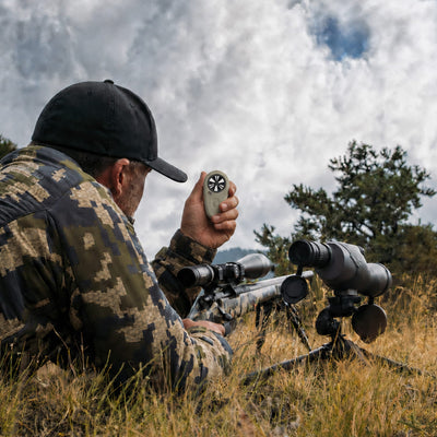 A man in camouflage clothing and a black cap lies in tall grass outdoors, holding a small handheld device near a rifle with a mounted scope. Another scope sits on a tripod nearby, with trees and a cloudy sky in the background.
